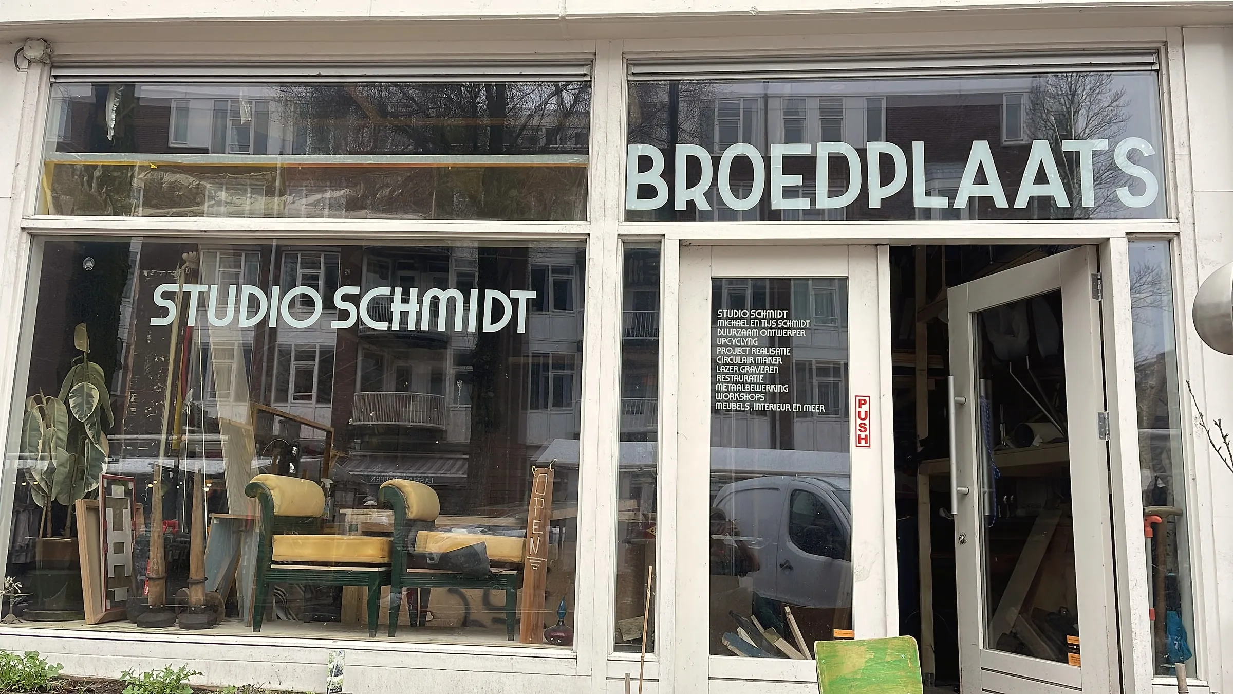 Winkelpui van Studio Schmidt aan de Dapperstraat: ramen met de letters STUDIO SCHMIDT en BROEDPLAATS, twee stoelen met groene frames en gele kussens in de etalage en de deur open